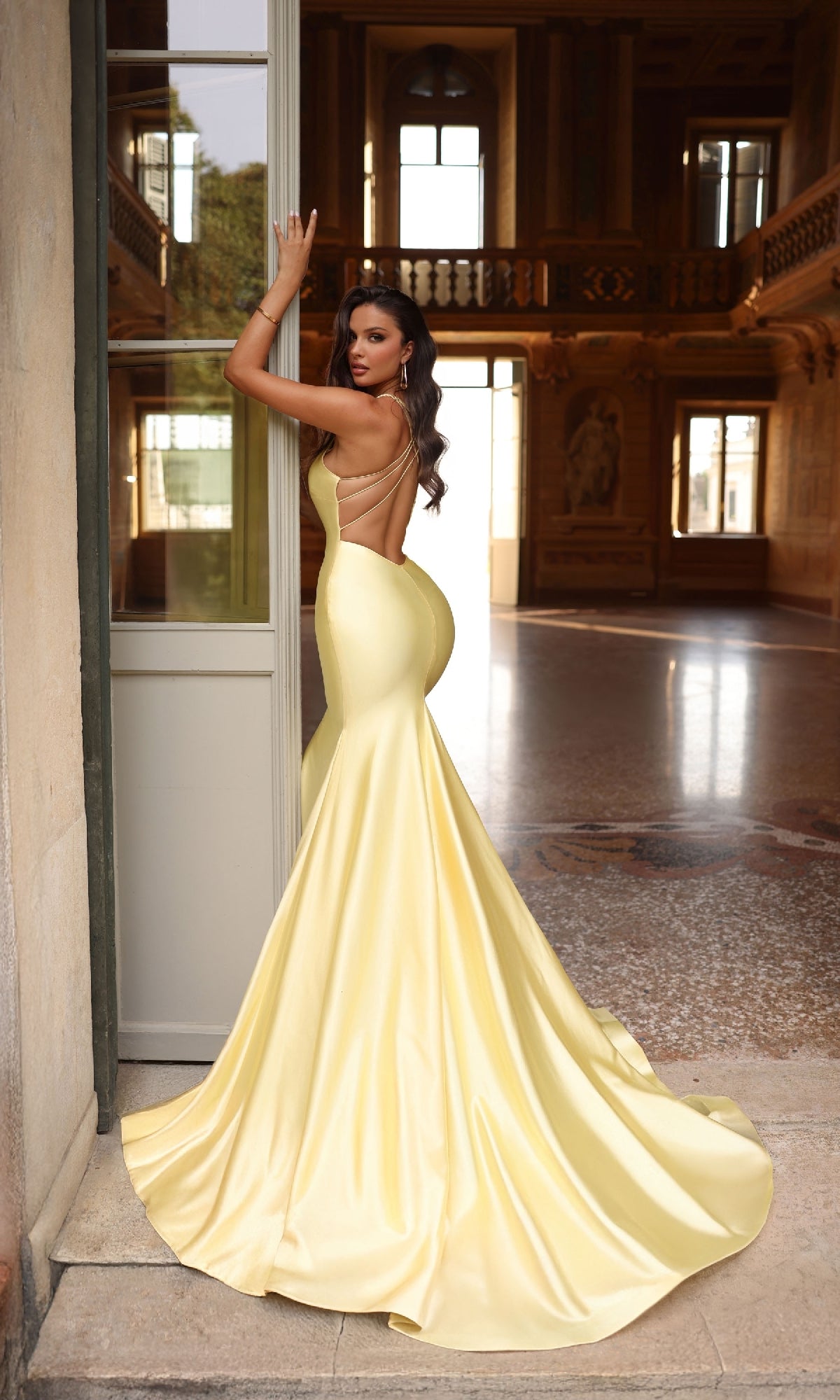 A woman wearing the Alyce 61966 long yellow satin prom dress with a backless design and v-neckline stands by a doorway in an ornate, high-ceilinged room with large windows.